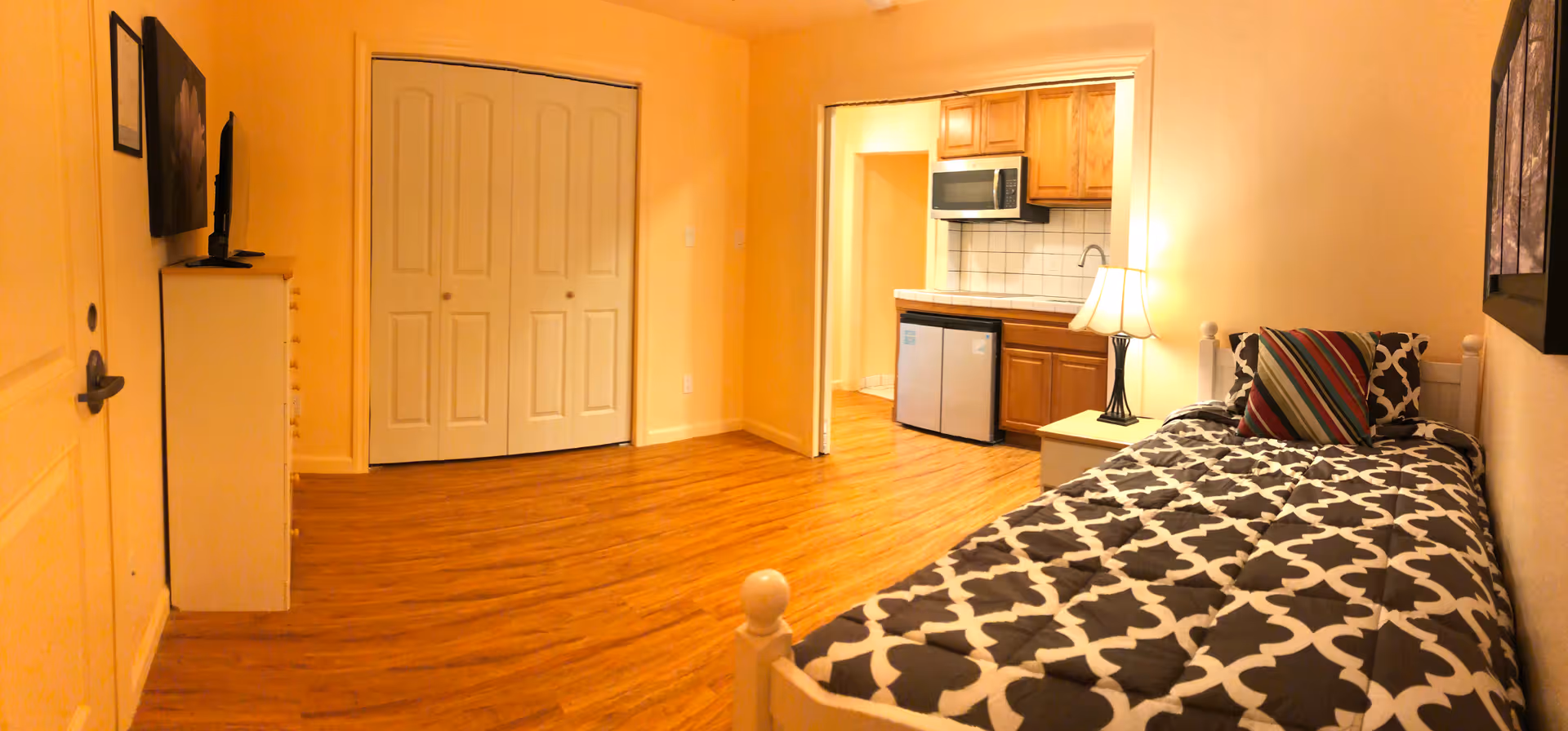 Studio-style bedroom with a single bed and bedside lamp, dresser with TV, closet doors, and a small kitchenette area.