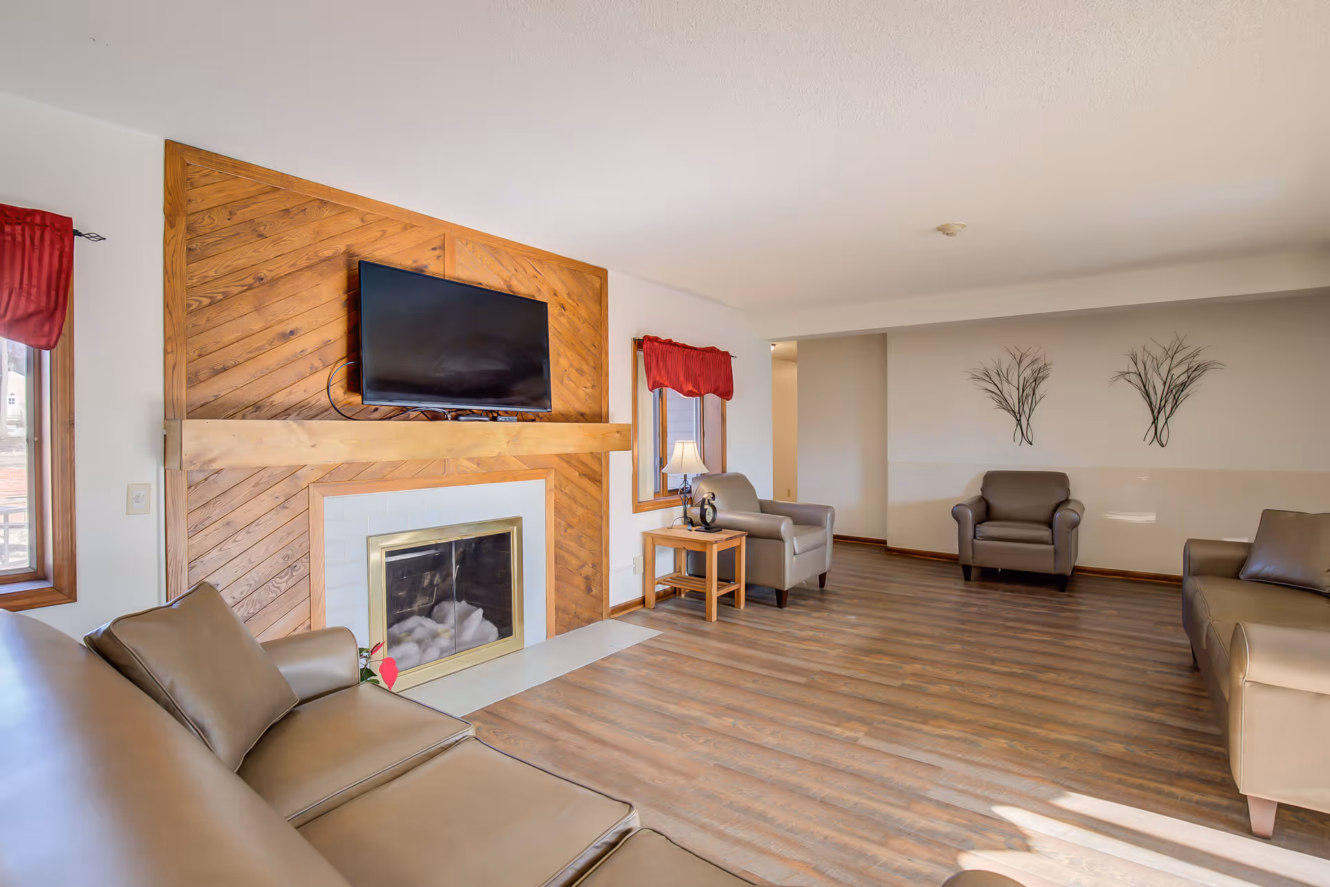 A spacious living room with wooden flooring, beige leather sofas and armchairs arranged around a fireplace with a wooden panel backdrop. A flat-screen TV is mounted above the fireplace. There are two windows with red curtains and a small wooden side table with a lamp. The walls are light-colored with decorative metal wall art.