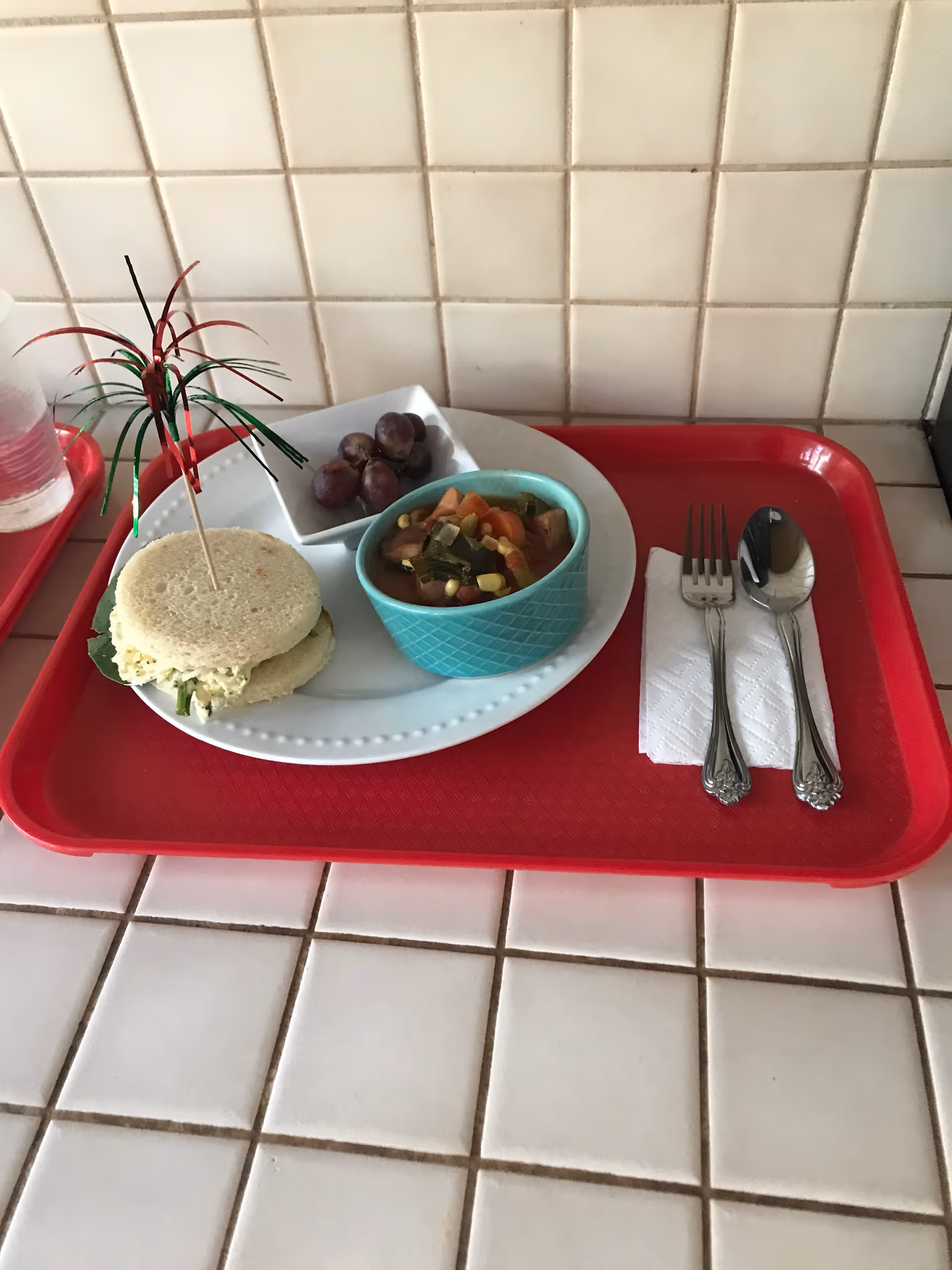 A red tray on a tiled countertop holds a white plate with a small sandwich decorated with a colorful cocktail pick, a small bowl of vegetable soup, and a small dish of grapes. Next to the plate are a fork and spoon placed on a folded white napkin.