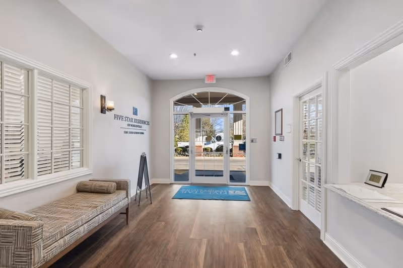 Entrance lobby of Five Star Residences of Noblesville featuring a patterned bench with a bolster pillow on the left, a blue welcome mat in front of glass double doors leading outside, and a reception counter on the right with a digital clock and papers.