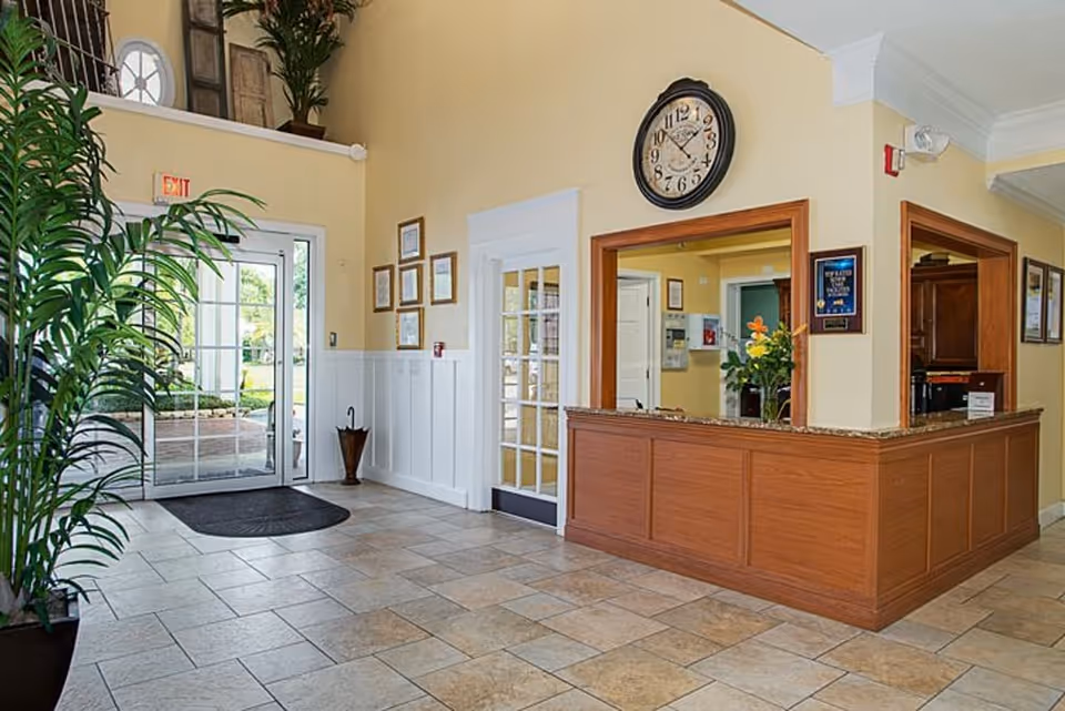 Reception area of a senior living facility with a wooden front desk, a large clock on the wall above the desk, framed certificates on the walls, a glass door entrance with an exit sign above it, and a tall green plant on the left side.