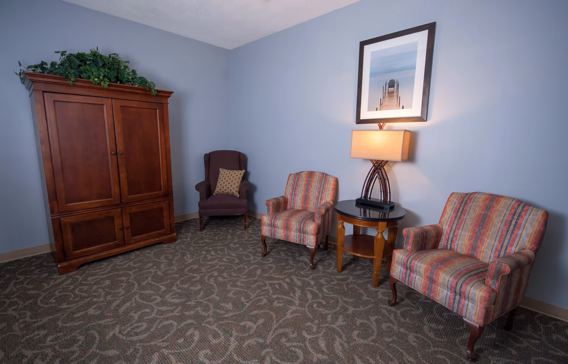 A cozy sitting area with two striped armchairs, a round wooden side table with a lamp, a framed picture of a pier on the wall, a dark wooden armoire with a green plant on top, and a purple wingback chair with a patterned pillow in the corner. The walls are painted light blue and the floor is covered with a patterned carpet.