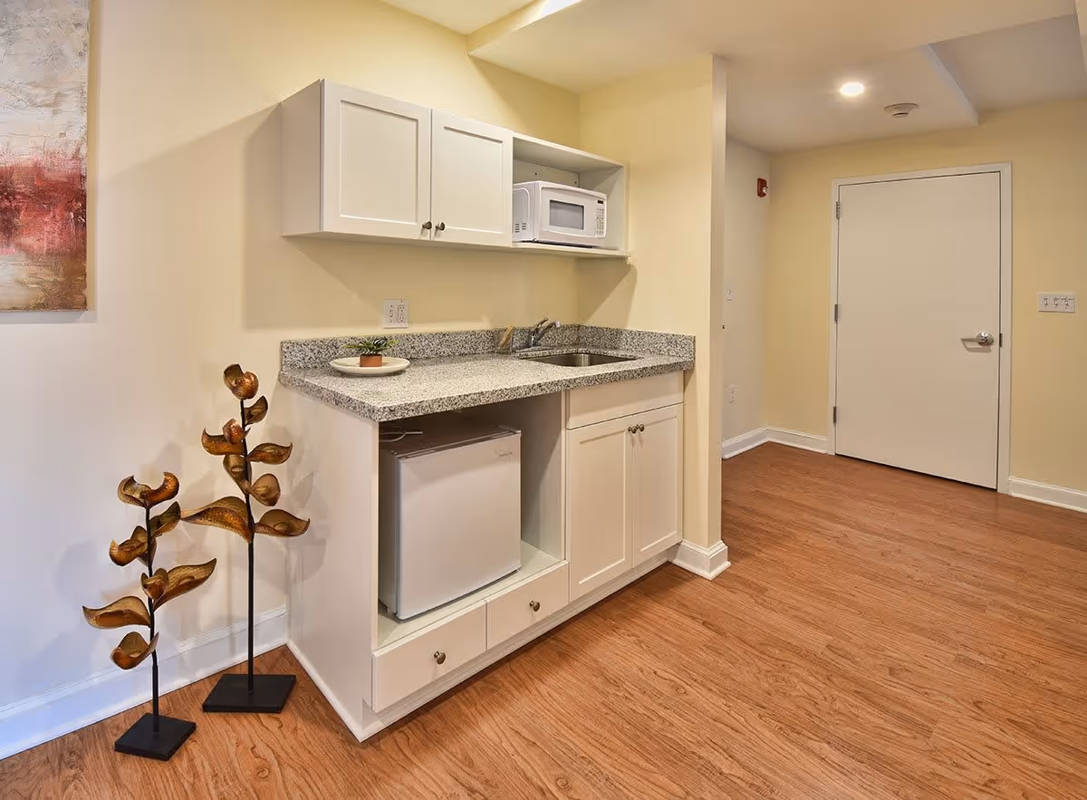 A small kitchenette area with white cabinets, a granite countertop, a microwave, a small refrigerator, and a sink. The floor is wooden, and there are two decorative metal sculptures resembling flowers on the left side. A closed white door is visible in the background.