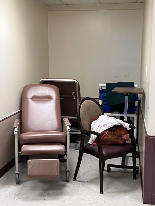 Narrow interior alcove storing a medical recliner, a guest chair piled with linens and a bag, and other medical equipment.