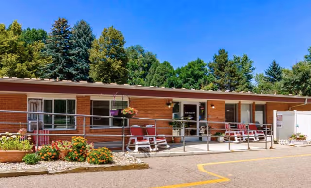 Single-story brick facility entrance with a wheelchair ramp, red chairs and potted plants outside under a blue sky and trees.