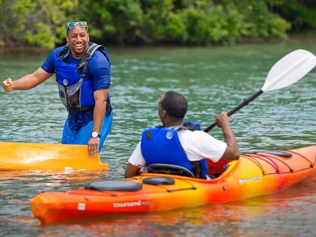 Two men enjoying kayaking on a calm body of water surrounded by greenery. One man is sitting in an orange kayak paddling, while the other man is standing in the water holding a yellow kayak and smiling. Both are wearing blue life jackets.