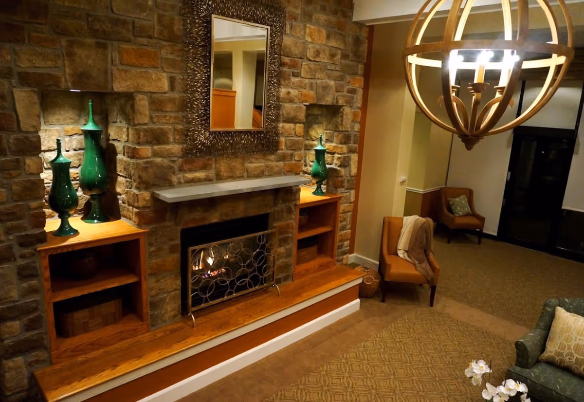 A cozy living room area with a stone fireplace featuring a lit fire, flanked by built-in wooden shelves holding green decorative vases. Above the fireplace is a decorative mirror with a textured frame. The room includes a brown armchair with a blanket draped over it, another armchair in the background, a patterned carpet, and a large spherical chandelier hanging from the ceiling.