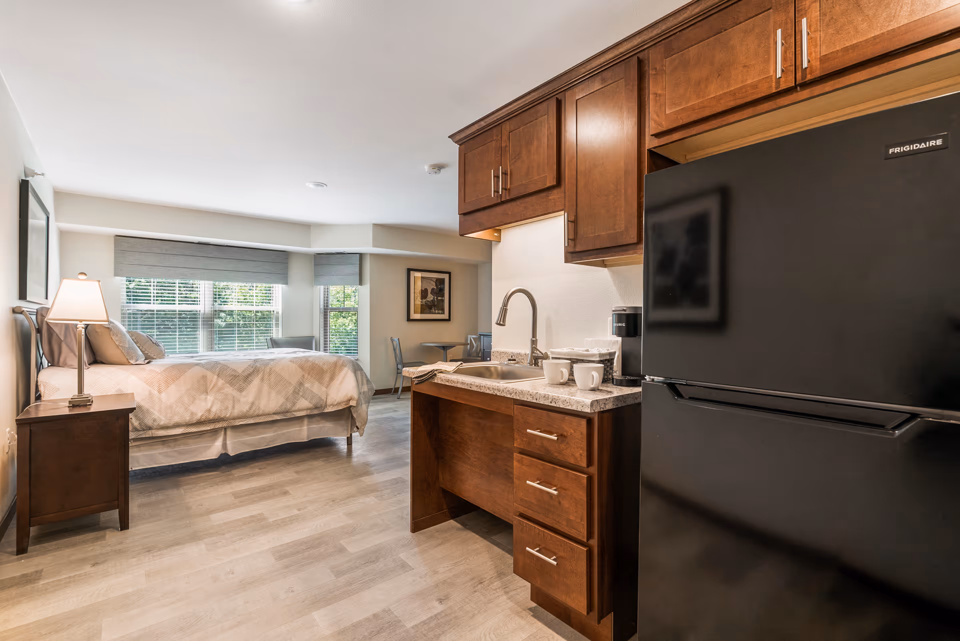 A senior living facility room at Cedar Creek Senior Living featuring a bed with a nightstand and lamp, a kitchenette with a sink, countertop, coffee maker, and a black refrigerator. There is a small dining table with two chairs near the window, and the room has large windows with blinds allowing natural light to enter.