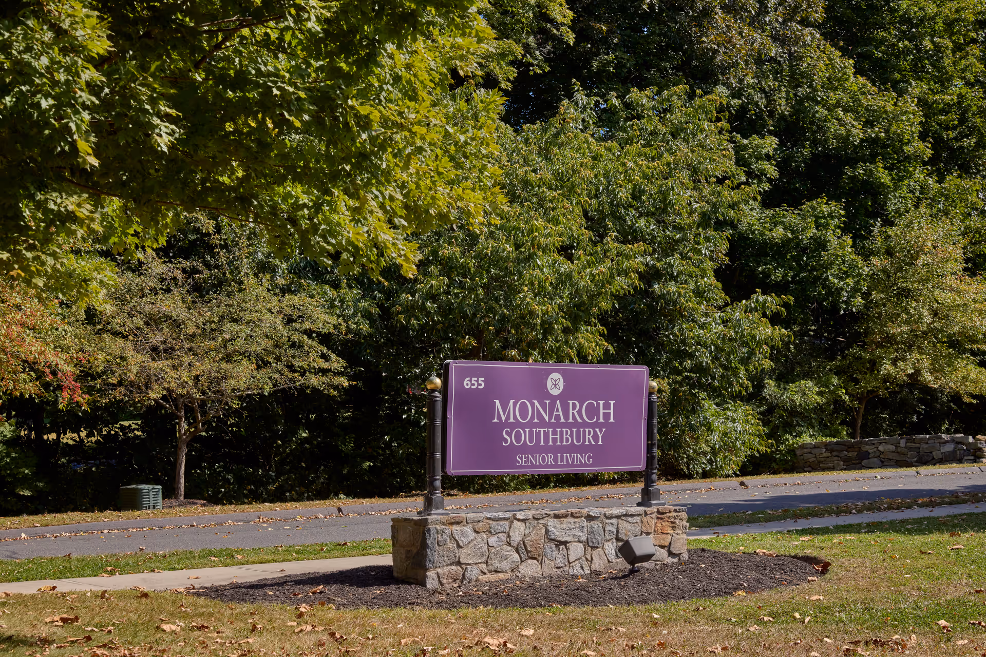 A purple sign with white text reading '655 Monarch Southbury Senior Living' mounted on a stone base, situated on a grassy area with trees and a road in the background.