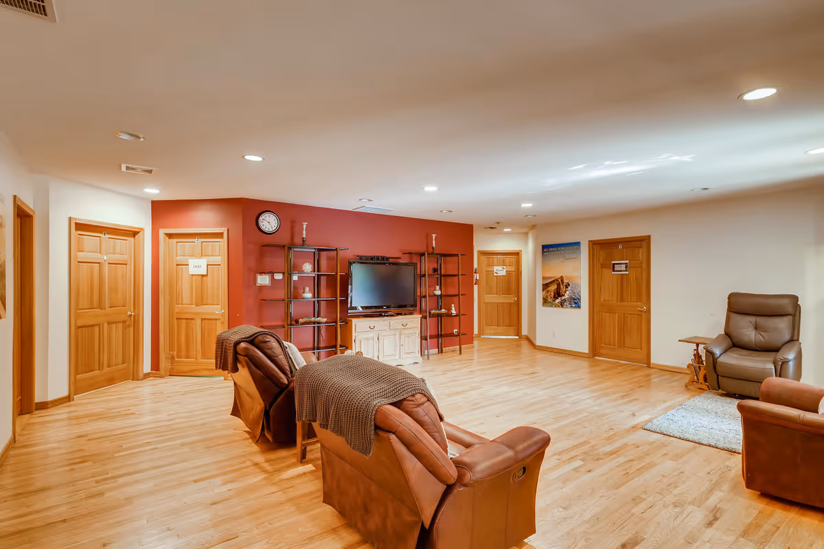 A spacious living room area with wooden flooring and multiple wooden doors along the walls. The room features two brown leather recliners with blankets draped over them, a large flat-screen TV mounted on a red accent wall with black metal shelves on either side, and a beige armchair with a small side table on a rug. The ceiling has recessed lighting, and there is a clock on the red wall above the TV.