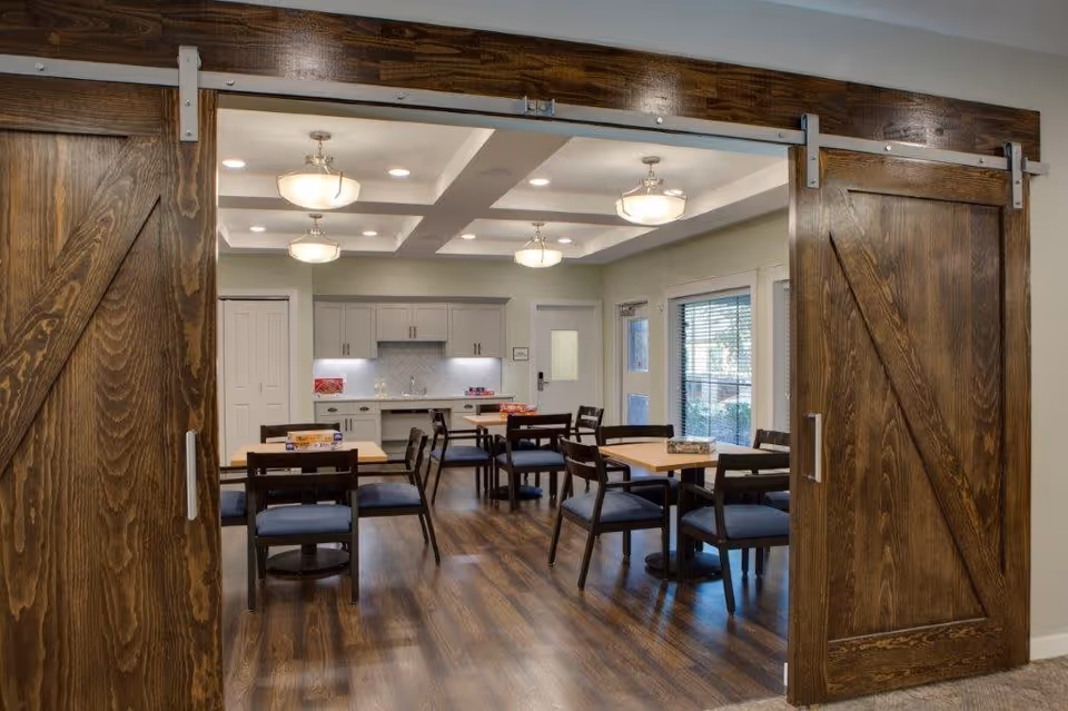 Interior view of a dining area in a senior living facility with wooden sliding barn doors, several tables and chairs, overhead lighting, and a kitchenette with cabinets and a countertop in the background.