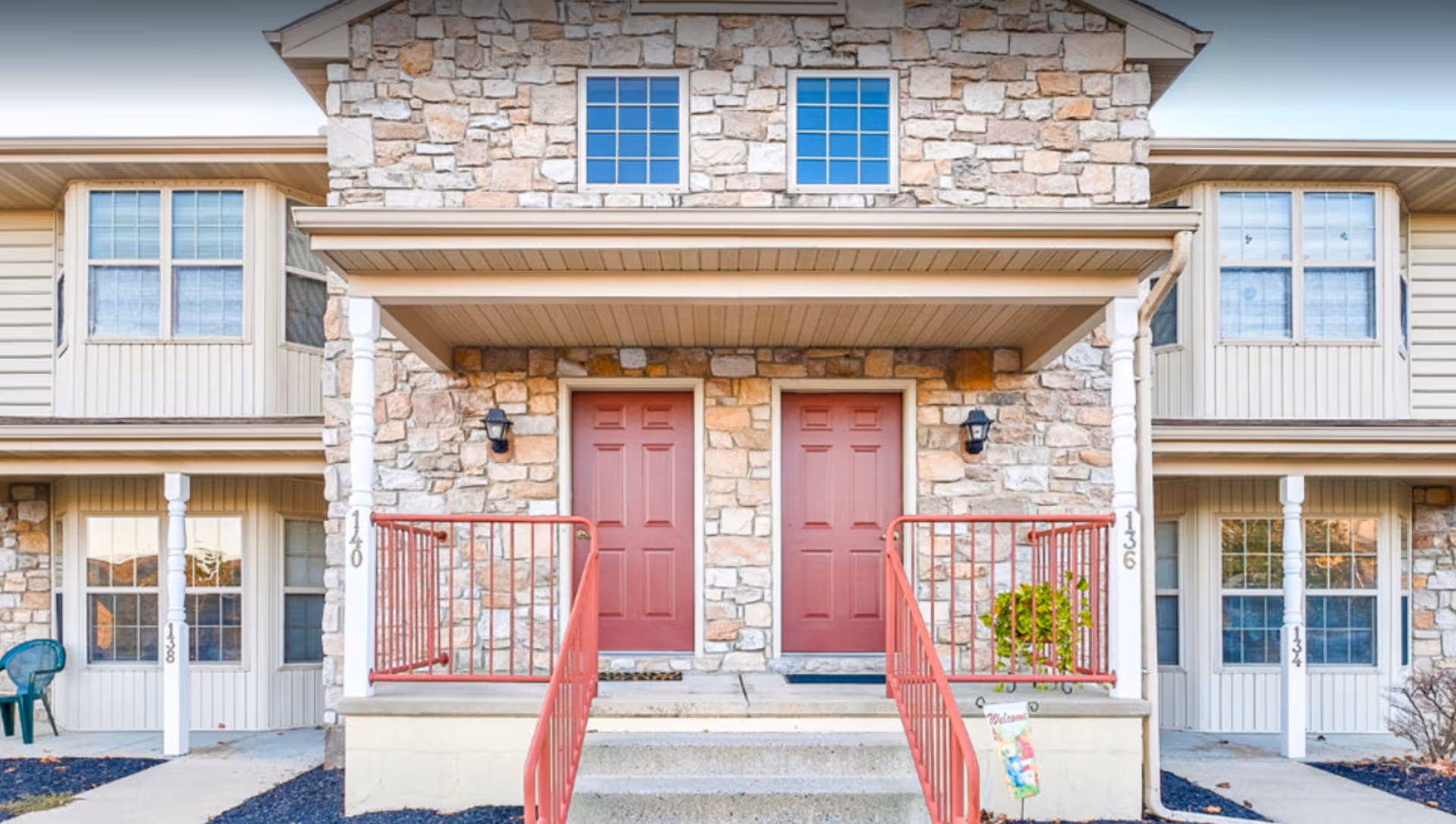 Front exterior view of a senior living facility building with stone and beige siding, featuring two red entrance doors with small porches and red railings, windows above and beside the doors, and a small welcome sign near the steps.