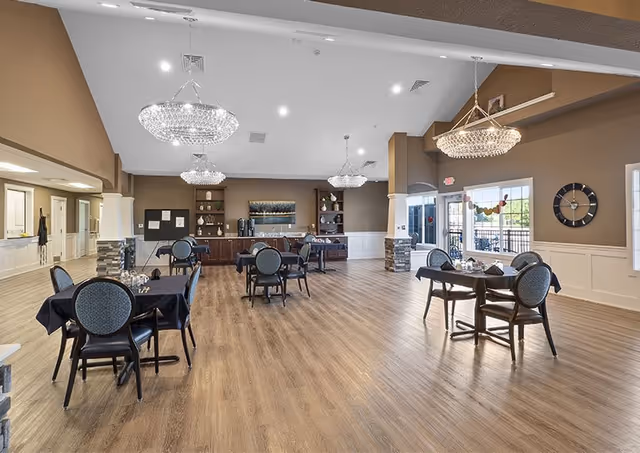 A spacious dining room with several tables covered in dark tablecloths and surrounded by chairs. The room features wooden flooring, large windows allowing natural light, and elegant chandeliers hanging from the ceiling. There is a counter area with coffee machines and shelves at the back of the room.