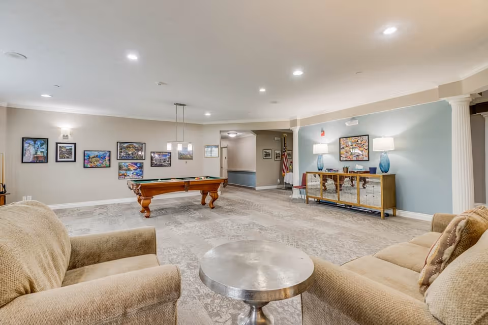 A spacious common area in an assisted living facility featuring a pool table in the center, beige armchairs with a round metallic table between them in the foreground, a sideboard with two blue lamps and a framed picture on a light blue wall, and several framed pictures on a beige wall. The room has recessed ceiling lights and carpeted flooring.