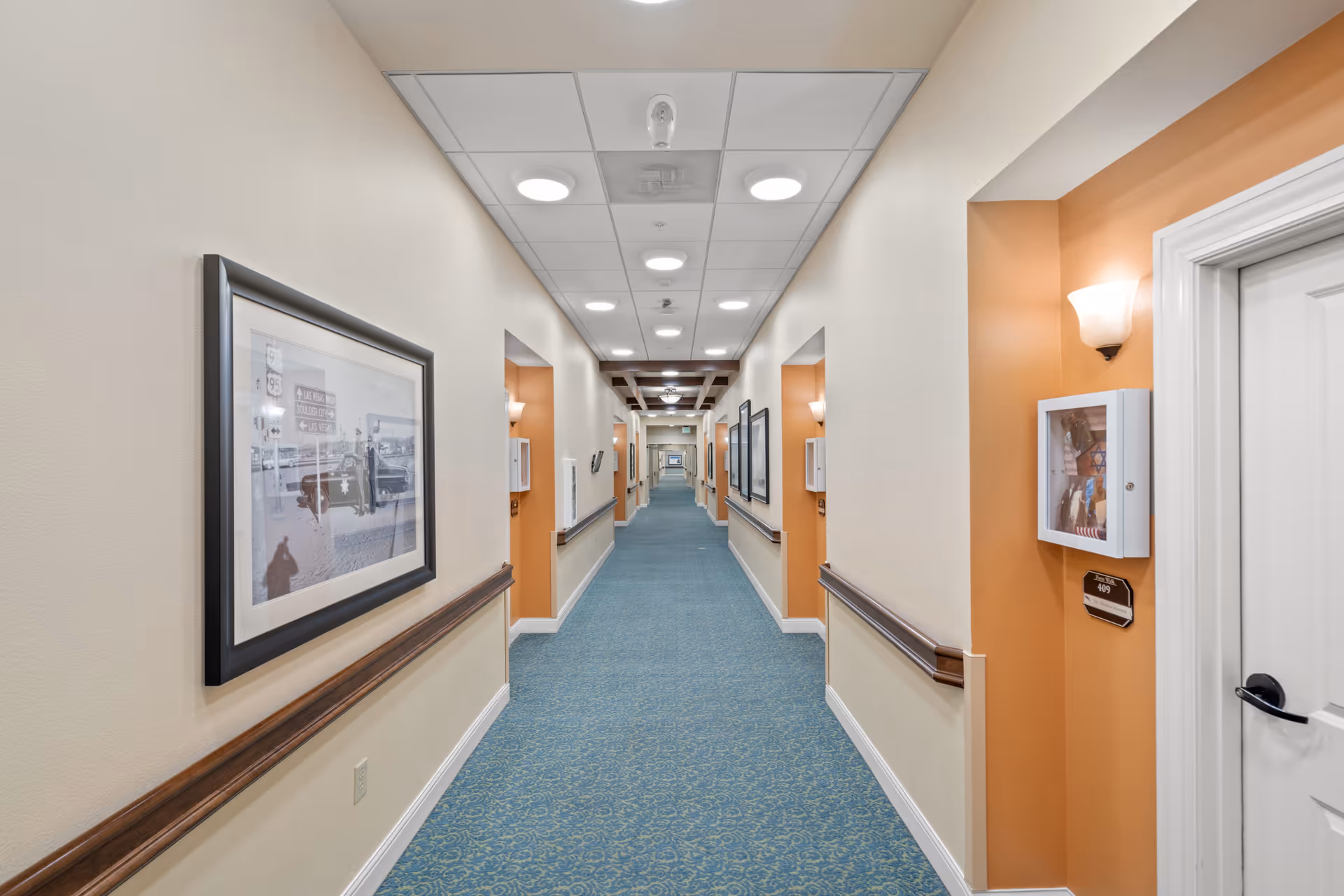 Long, well-lit hallway in a senior living facility with blue patterned carpet, beige and orange walls, framed pictures on the walls, handrails along both sides, and white doors leading to rooms.