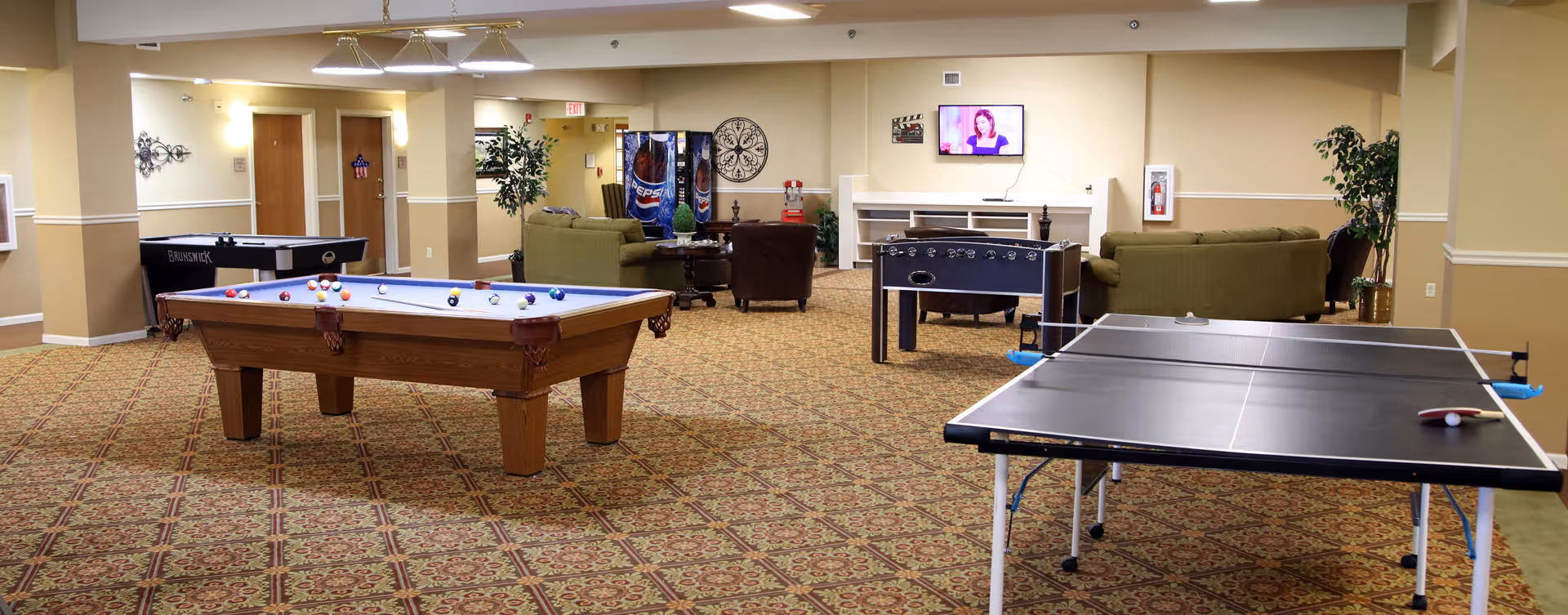 A spacious recreational room with a pool table, ping pong table, foosball table, and a shuffleboard table. There are several green sofas and chairs arranged around a TV mounted on the wall. The room has patterned carpet flooring, beige walls, and some potted plants for decoration. A Pepsi vending machine is visible in the background.