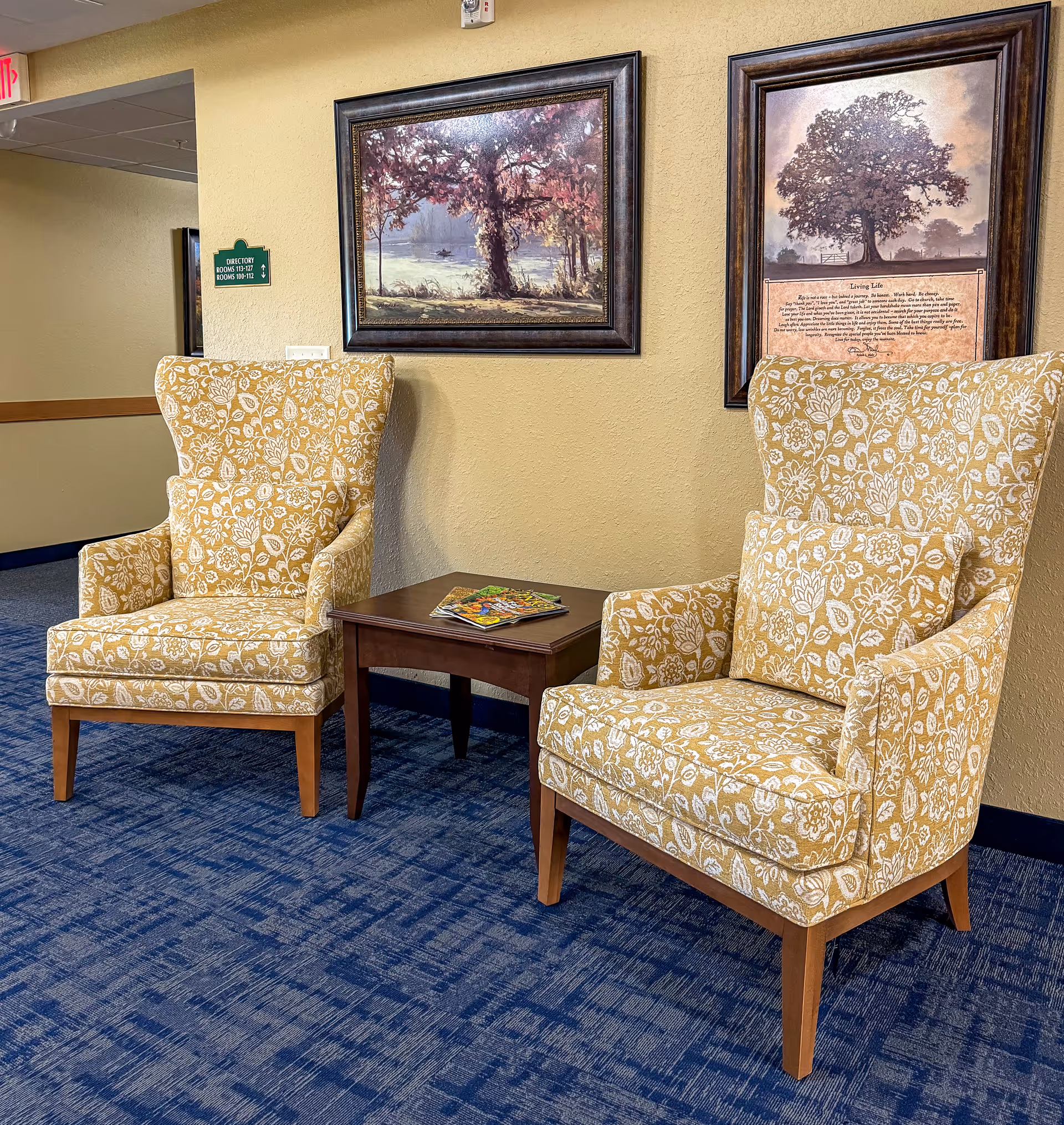 Two yellow floral patterned armchairs with matching cushions are placed on a blue carpeted floor with a small wooden table between them holding magazines. Behind the chairs, there are two framed pictures hanging on a beige wall. A green directory sign is visible on the left side of the wall.