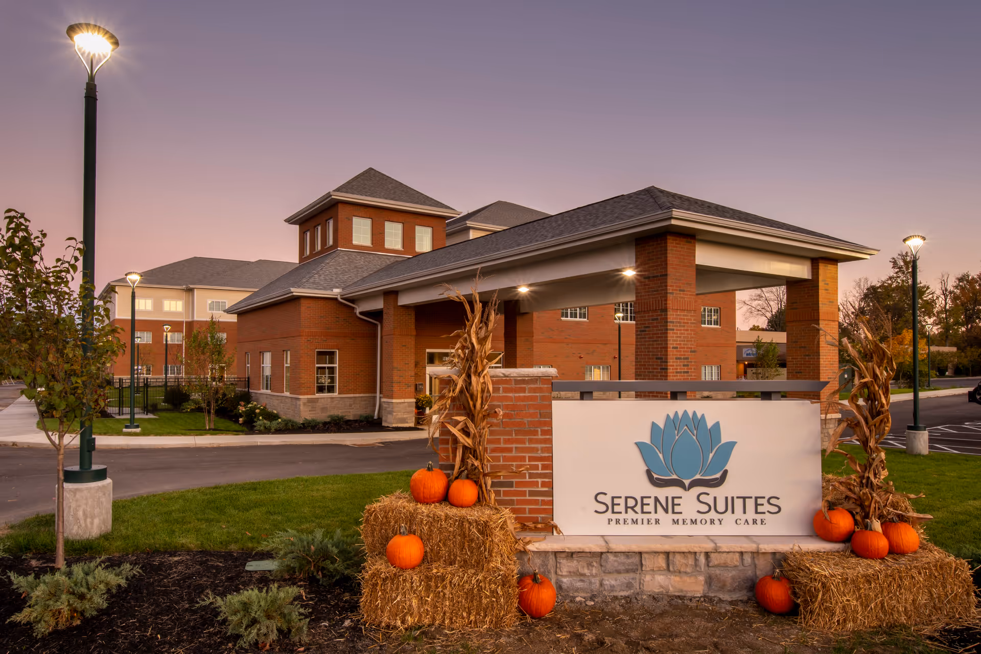 Exterior view of Serene Suites, a brick building with a covered entrance and multiple windows, surrounded by a landscaped area with trees and street lamps at dusk. In front, there is a sign with the Serene Suites logo and text, decorated with hay bales, dried corn stalks, and orange pumpkins.