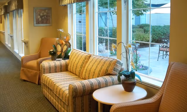 A cozy seating area with a striped sofa, armchairs, and potted plants facing large windows that look out onto a courtyard.