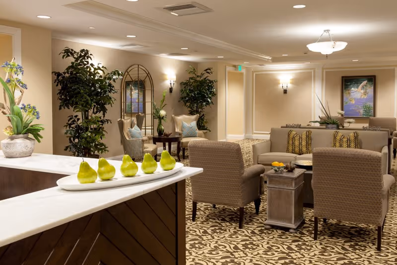 A well-lit, elegant common area with patterned carpet, beige walls, and comfortable seating including armchairs and sofas arranged around small tables. Decorative plants and artwork adorn the space, and a white countertop with a tray of green pears is visible in the foreground.
