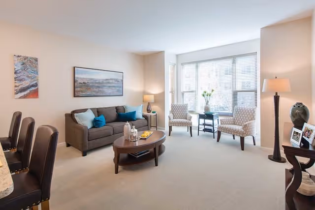 A bright furnished living room with a sofa, coffee table, two armchairs by a large window, and lamps.