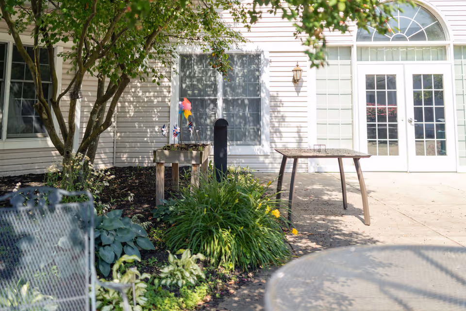 Outdoor patio area with a garden bed containing green plants and flowers, a small wooden planter with colorful pinwheels, a metal mesh chair, a table with four legs, and a building with white siding and large windows and glass doors in the background.