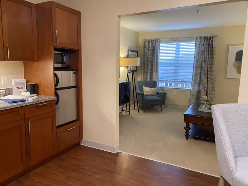 Interior view of a senior living facility apartment showing a small kitchen area with wooden cabinets, a microwave, and a mini refrigerator on the left. The kitchen opens into a living room area with a blue armchair, a floor lamp, a coffee table, and a window with patterned curtains letting in natural light.