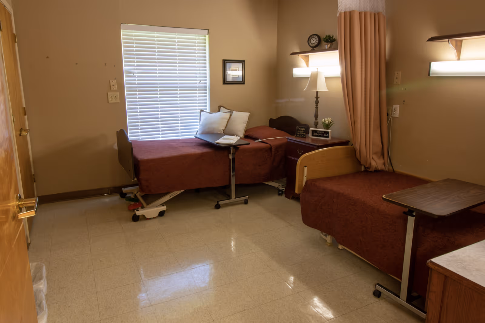 A shared senior room with two medical-style twin beds covered in burgundy blankets, a window with blinds, a nightstand and lamp, and overbed tables.