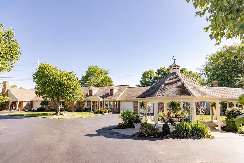 Exterior view of Westover Retirement Community showing a single-story brick building with a pitched roof and dormer windows. In the foreground, there is a gazebo with a weather vane on top, surrounded by greenery and plants. Two people are seated under the gazebo, enjoying the outdoor space on a sunny day with clear blue skies.