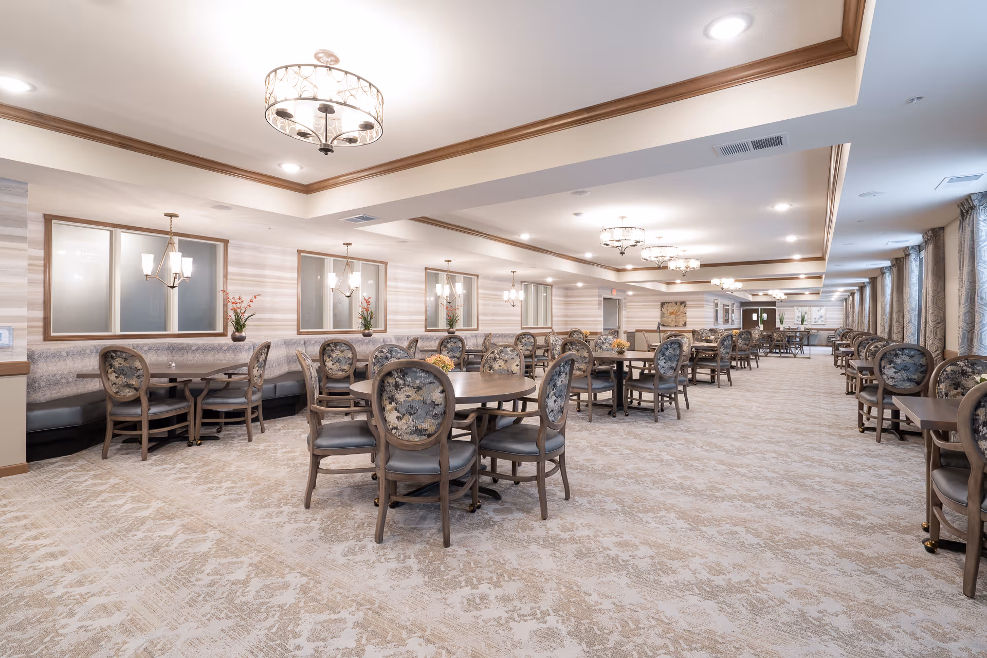 A spacious and well-lit dining room in a senior living facility with multiple round and square tables surrounded by floral upholstered chairs. The room features elegant light fixtures hanging from a tray ceiling, large windows with curtains along one side, and decorative wall sconces with flowers on the walls.