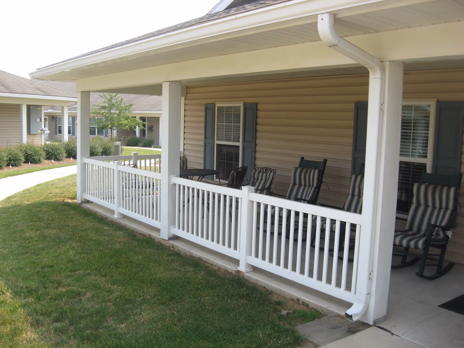 Covered porch area with white railing and several cushioned rocking chairs with striped cushions. The porch is attached to a beige building with blue shutters on the windows. There is a small round table with chairs on the porch. Green grass and a paved walkway are visible in the foreground and background.