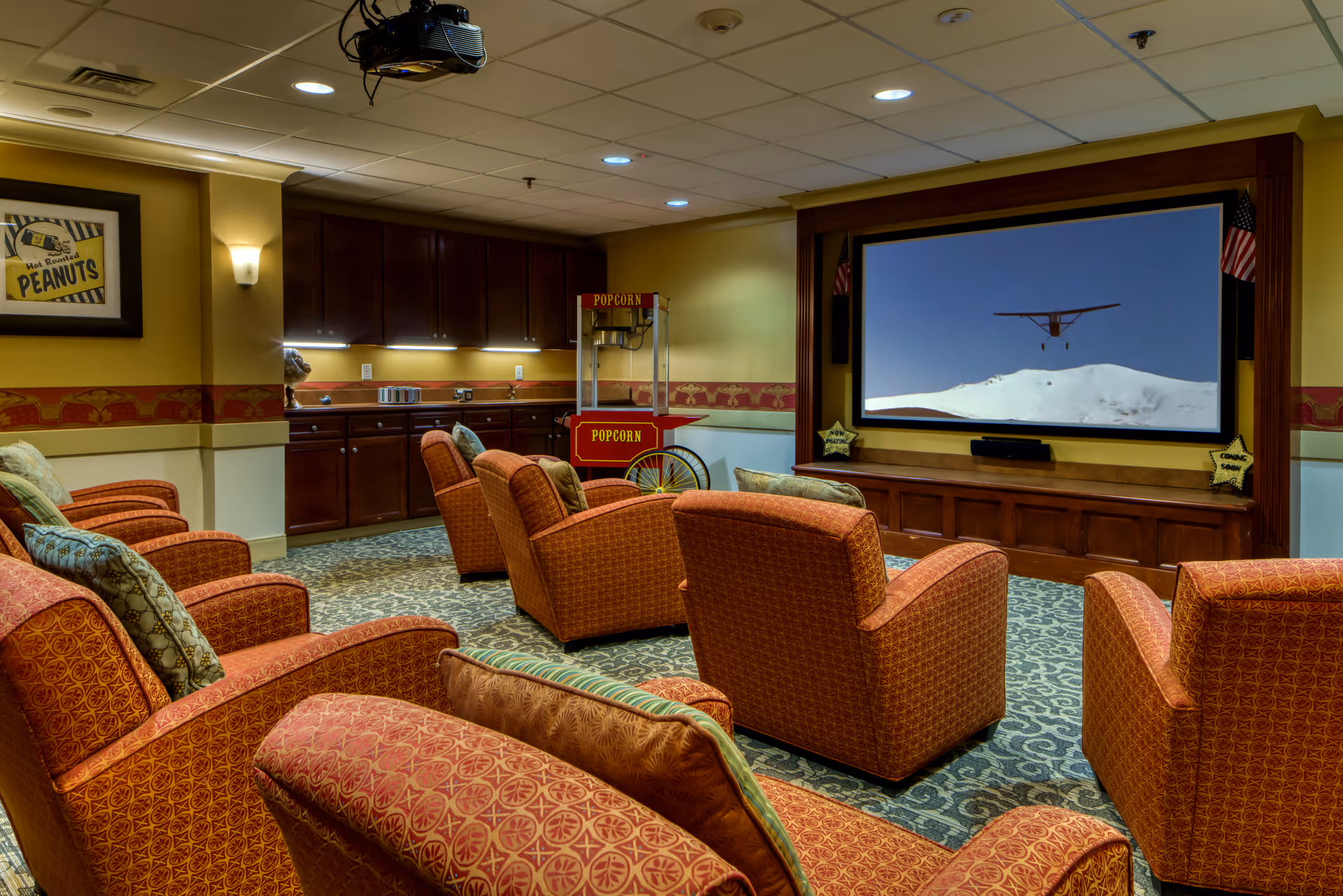 A cozy movie theater room with multiple cushioned armchairs arranged in rows facing a large screen showing a small airplane flying over snowy hills. There is a popcorn machine near the back wall, which also features dark wooden cabinets and a countertop. The room has warm yellow walls with decorative trim and a framed picture that reads 'Hot Roasted Peanuts'.