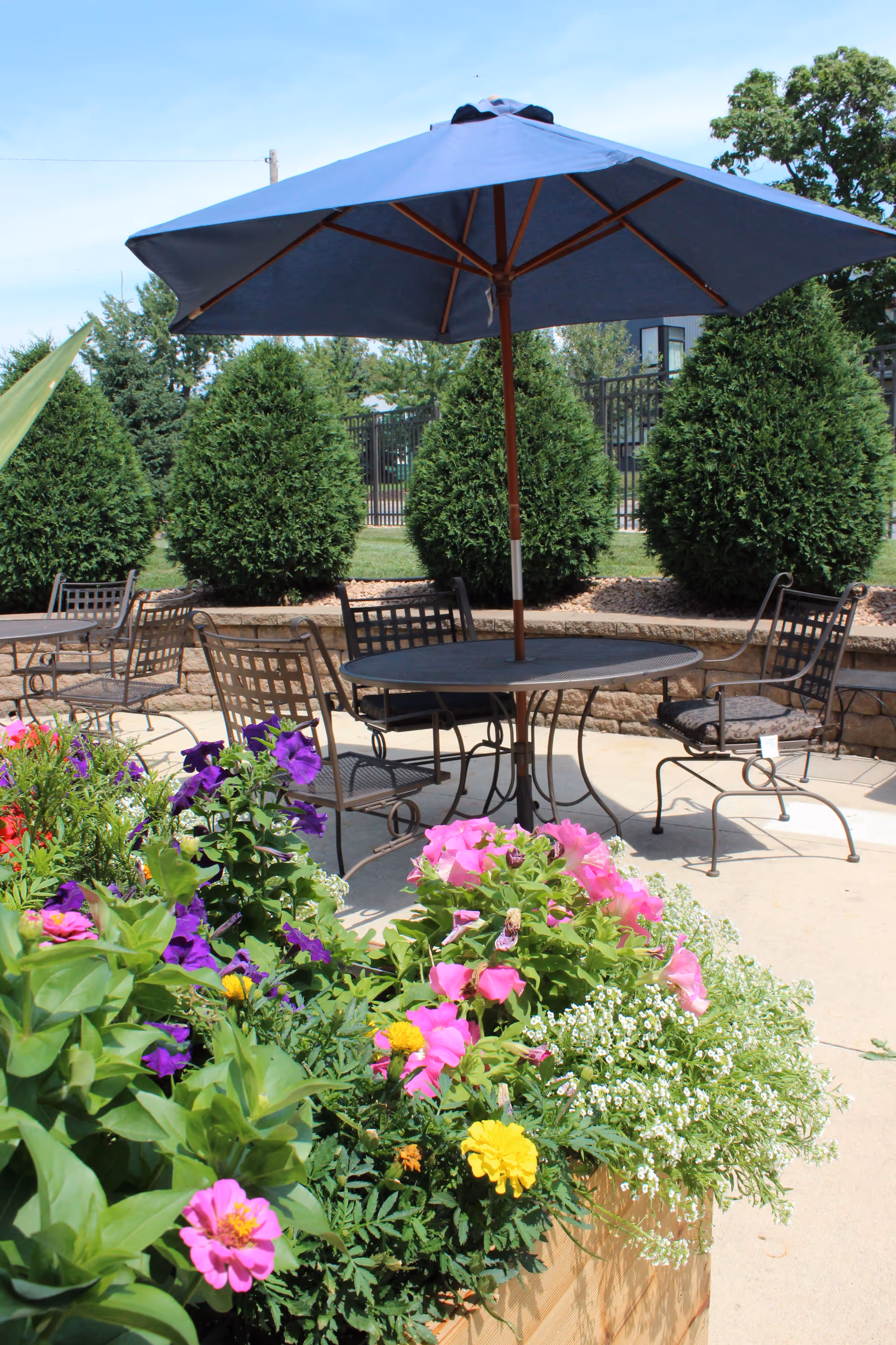 Outdoor patio area with metal tables and chairs under a large blue umbrella. In the foreground, there is a planter box filled with colorful flowers including pink, purple, yellow, and white blooms. Behind the patio, there are neatly trimmed evergreen bushes and a fence under a clear blue sky.