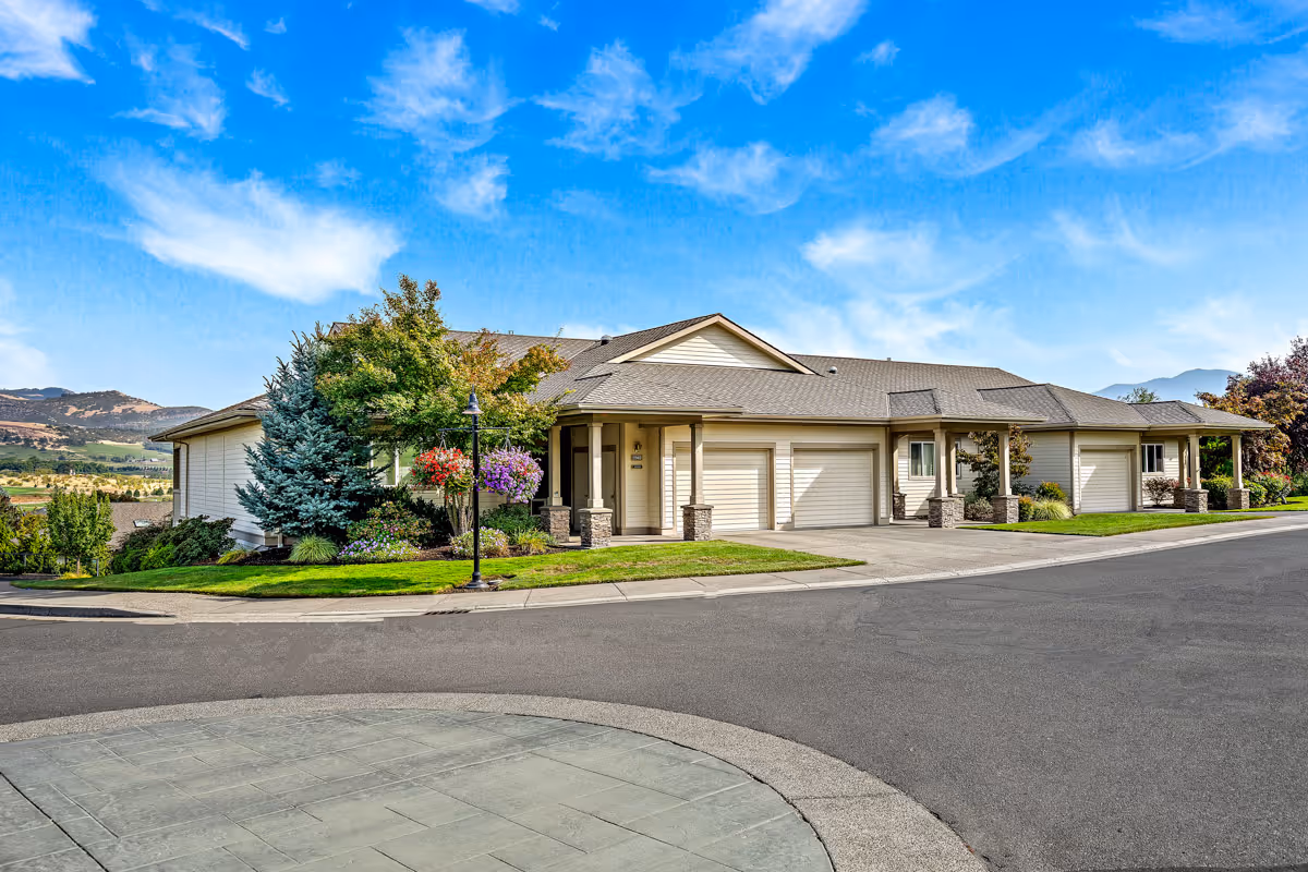 Single-story senior living building with attached garages, landscaped front yard, a lamppost, and mountains under a blue sky.