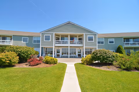 Front exterior of a two-story senior living building with a central covered entrance, balconies, and manicured lawn under a clear blue sky.