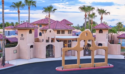 Exterior view of Arbor Ridge senior living facility featuring southwestern-style architecture with beige stucco walls, red tile roofs, palm trees, and a decorative entrance structure with the address number 9257.