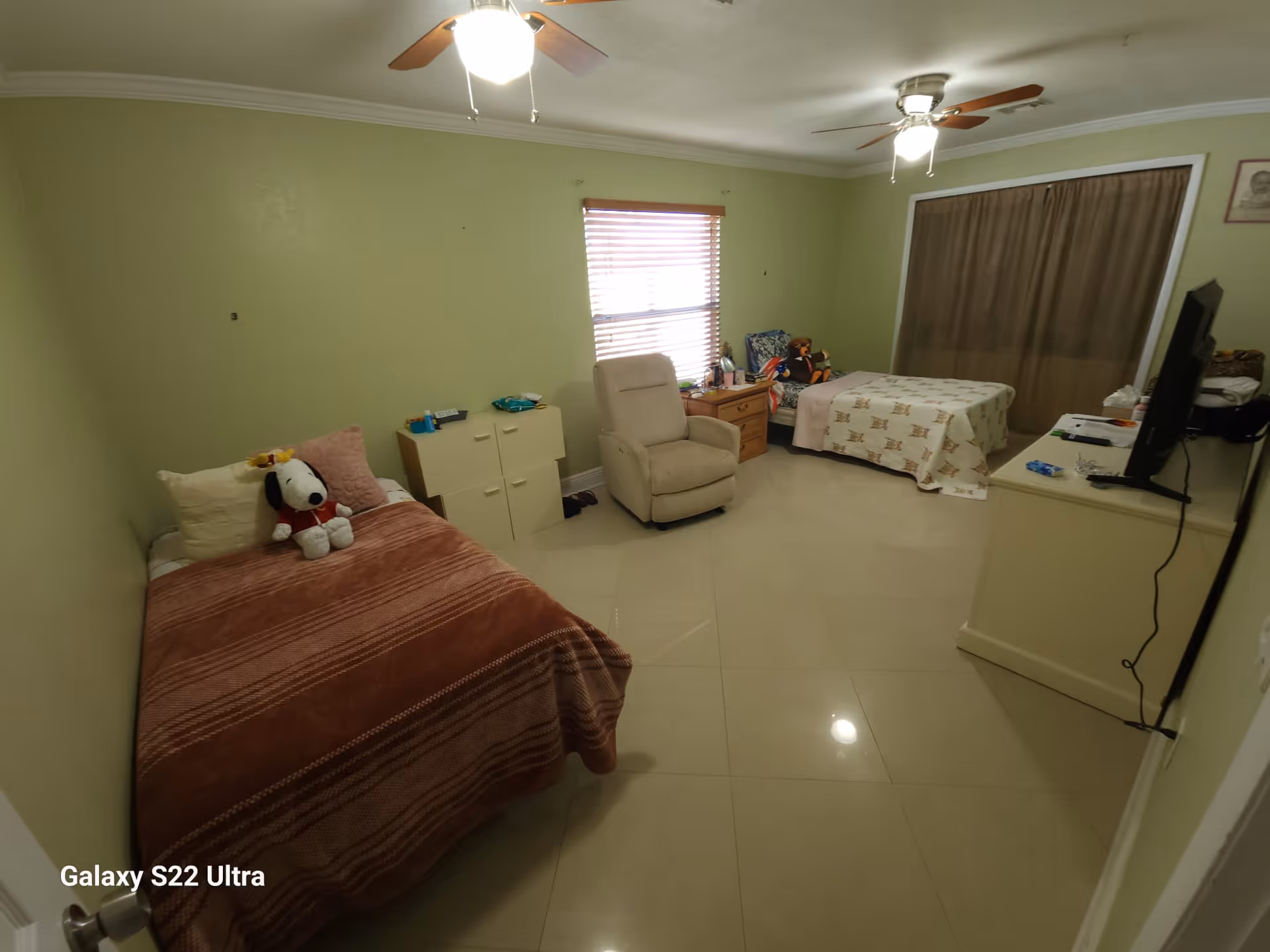 A bedroom in an assisted living facility with two single beds, one on the left with a brown blanket and a Snoopy stuffed toy, and one on the right with a patterned blanket. There is a beige recliner chair between the beds, a small cabinet, a wooden nightstand with items on it, and a TV on a dresser on the right side. The room has light green walls, a window with blinds, and a ceiling fan with a light.
