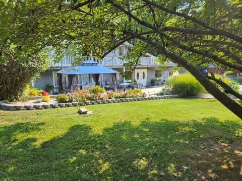 A sunny outdoor garden area with a large tree providing shade over a green lawn. In the background, there is a building with a covered patio featuring a blue canopy and outdoor seating surrounded by plants and flowers.