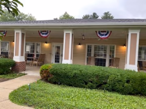 Exterior view of a single-story building with a covered porch featuring white columns and beige siding. The porch has rocking chairs and small tables, with patriotic red, white, and blue bunting decorations hanging above. There are well-maintained green bushes and a walkway leading to the porch.