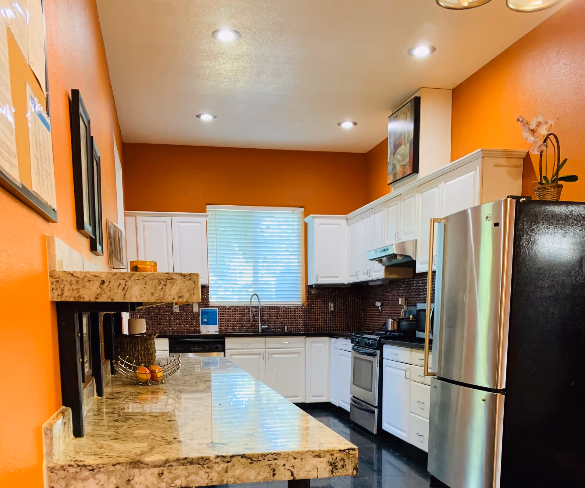 A modern kitchen with orange walls, white cabinets, and a granite countertop. The kitchen features a stainless steel refrigerator, a stove with an overhead vent, a dishwasher, and a window with blinds letting in natural light. There are framed pictures on the left wall and a small plant on top of the refrigerator.