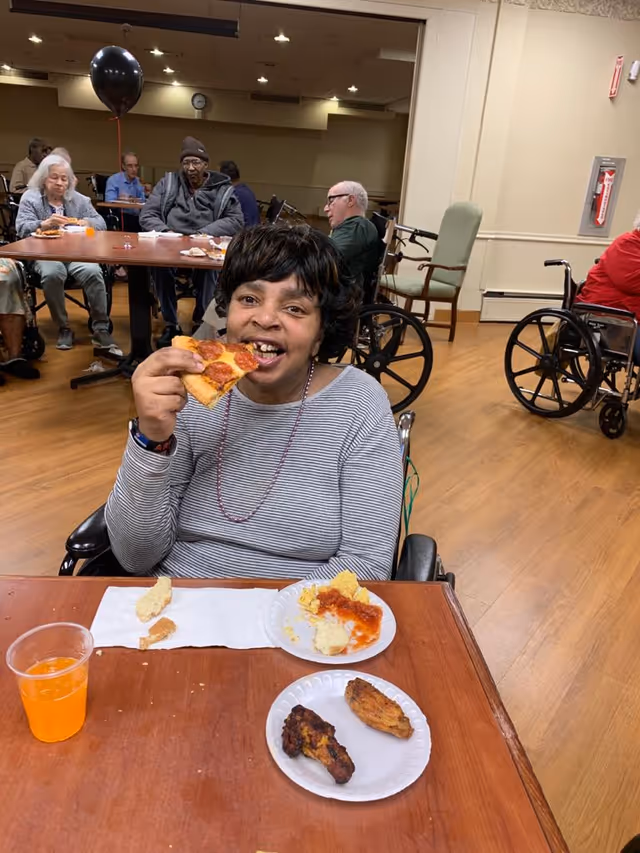 A woman in a wheelchair eating a slice of pepperoni pizza at a wooden table with plates of food and a cup of orange drink. In the background, several elderly people are seated at tables in a communal dining area, some in wheelchairs.