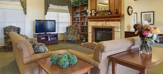 A cozy living room area with two beige sofas facing each other, a wooden coffee table with decorative green spheres, a flat-screen TV on a wooden stand, two patterned armchairs, a stone fireplace with a wooden mantel, built-in bookshelves, and a side table with a vase of flowers.