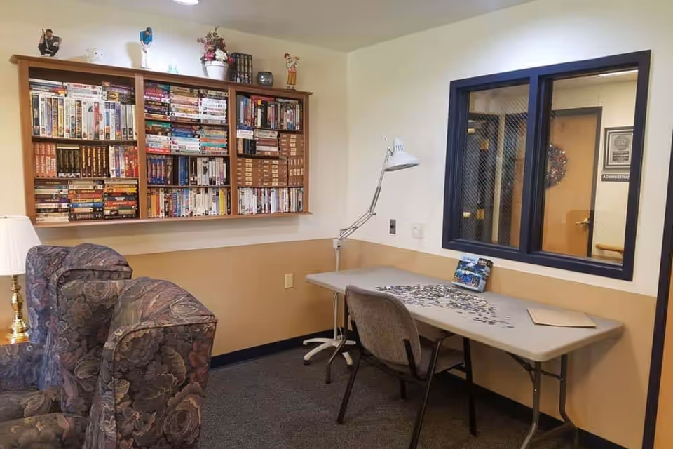 A cozy room with a wall-mounted bookshelf filled with books and DVDs, two patterned armchairs, a table with a puzzle in progress, a chair, and a desk lamp. There is a window with a view into another room labeled 'Administration'.