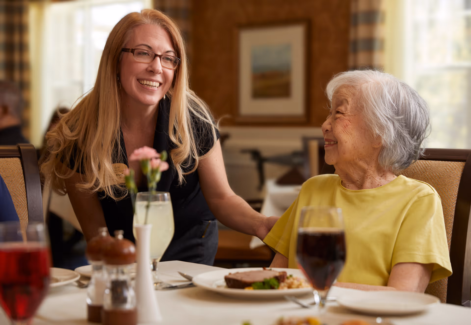 A smiling woman with long blonde hair and glasses leans toward an elderly woman with short gray hair wearing a yellow shirt, seated at a dining table with plates of food and drinks in a warmly lit room.