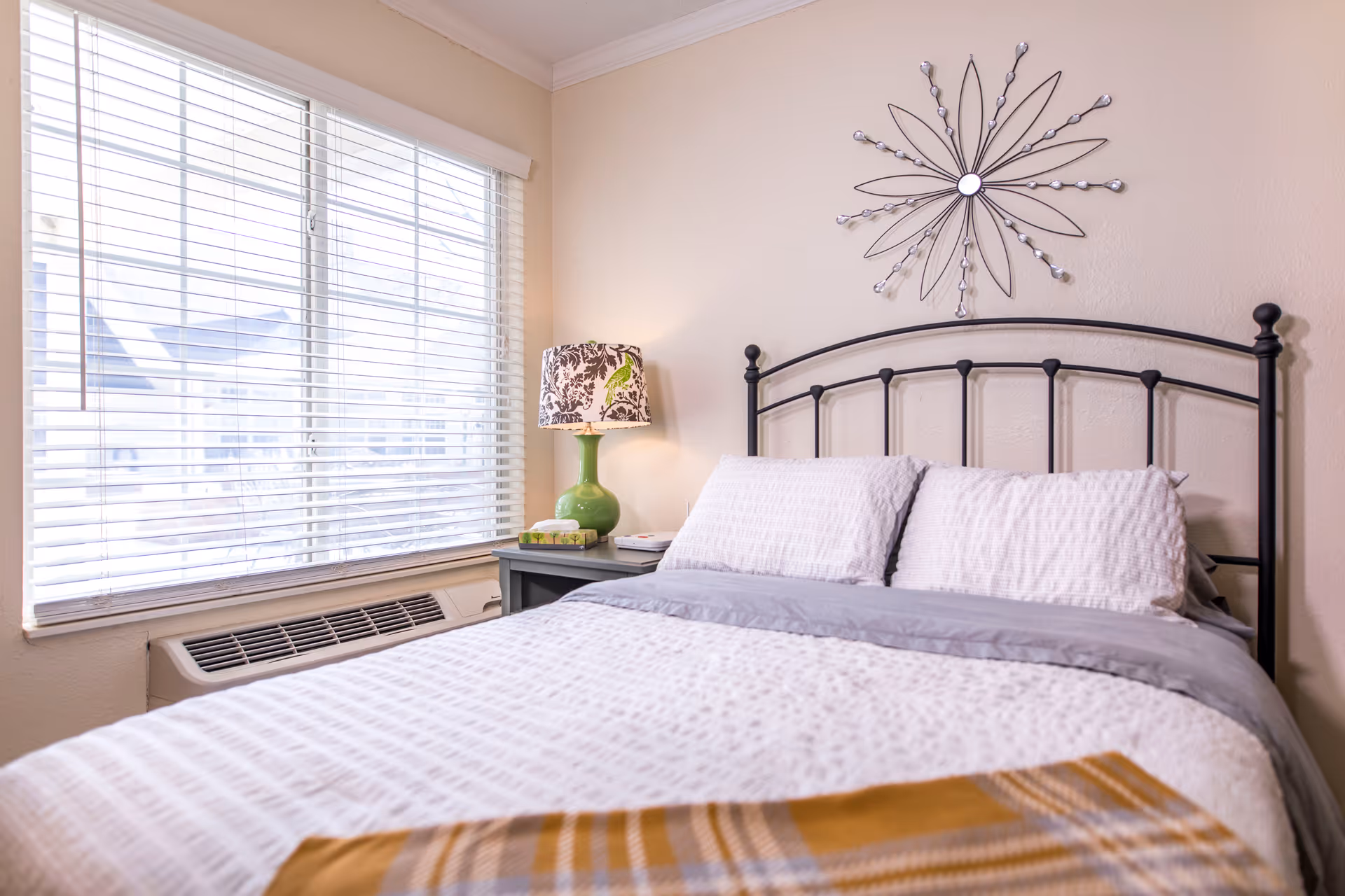 Well-lit bedroom with a made bed, metal headboard, bedside table with lamp, and a large window with blinds.