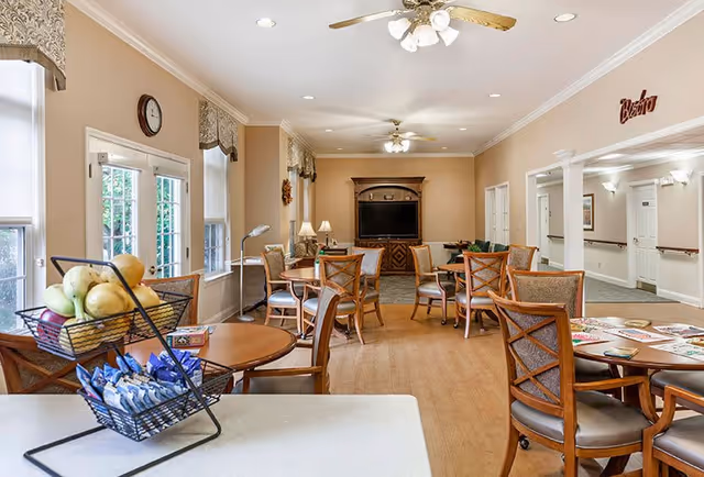 A bright and spacious common area with multiple round wooden tables and chairs arranged for seating. There is a basket of fruit and snacks on a counter in the foreground. The room features large windows with valances, ceiling fans with lights, and a wooden entertainment center with a TV at the far end. The walls are painted beige, and there is a sign that reads 'Bistro' above an open hallway.