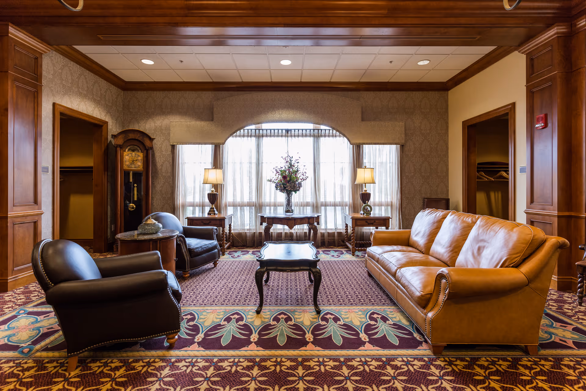 A cozy living room area with a brown leather sofa, two black leather armchairs, a wooden coffee table, and side tables with lamps. The room features patterned carpet, wood-paneled walls, a large window with sheer curtains, and a grandfather clock in the corner.