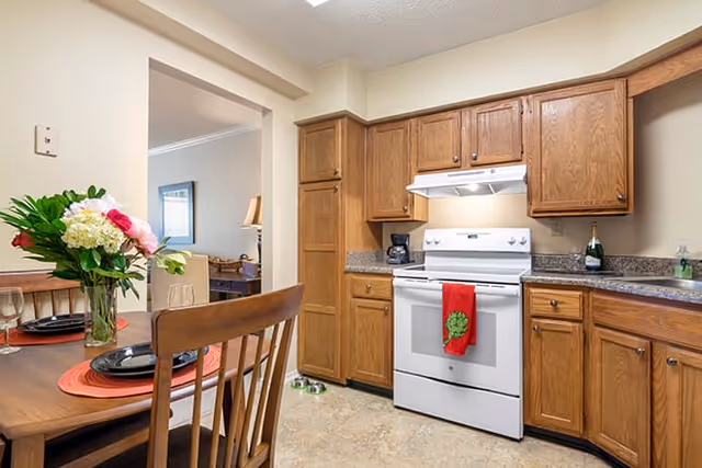 A kitchen with wooden cabinets, a white electric stove with a red towel hanging on the handle, a coffee maker on the counter, and a dining table set with plates, glasses, and a vase of flowers. The kitchen opens into a living area with a lamp and framed artwork visible.
