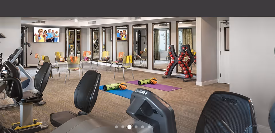 Bright fitness room with exercise bikes, yoga mats, dumbbells, chairs, and TVs in a senior living facility.