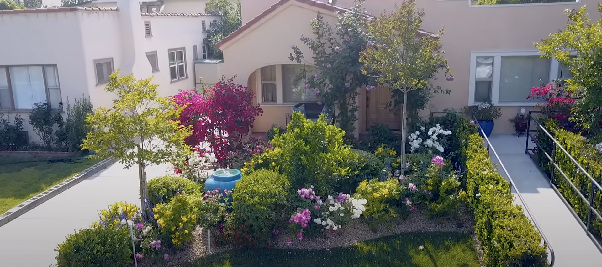 Front exterior view of a residential building with a well-maintained garden featuring various colorful flowers, shrubs, and small trees. There is a concrete pathway leading to the front door and a wheelchair accessible ramp on the right side.